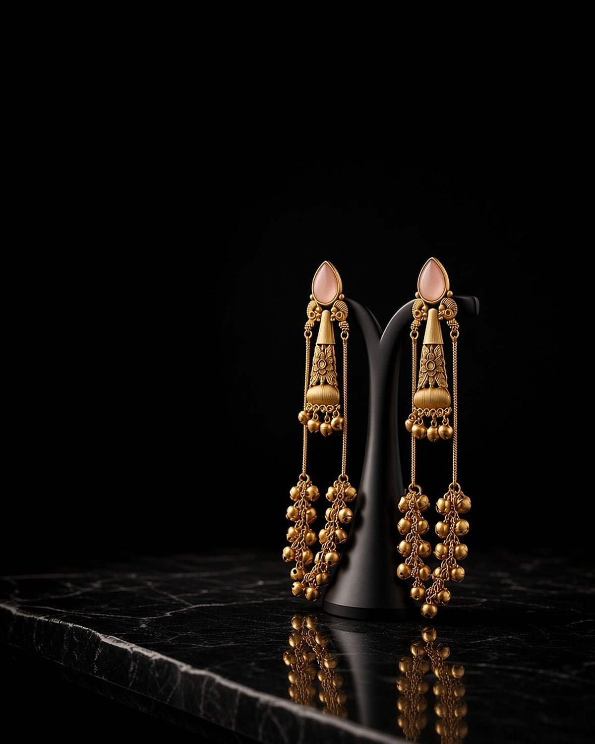 Gold ghungroo jhumka drop earrings with pink stones on a black stand against a dark background